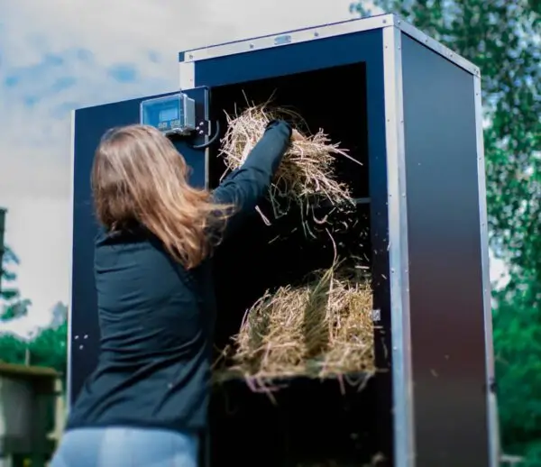 En stor box som är en höautomat som står vid en hästhage. En kvinna fyller höboxen med hästhö